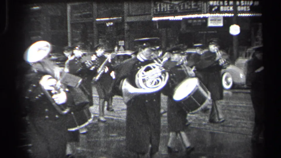 1937 Cleveland marching band frame with a drummer, brass players, wet pavement, and a theatre marquee in the background.
