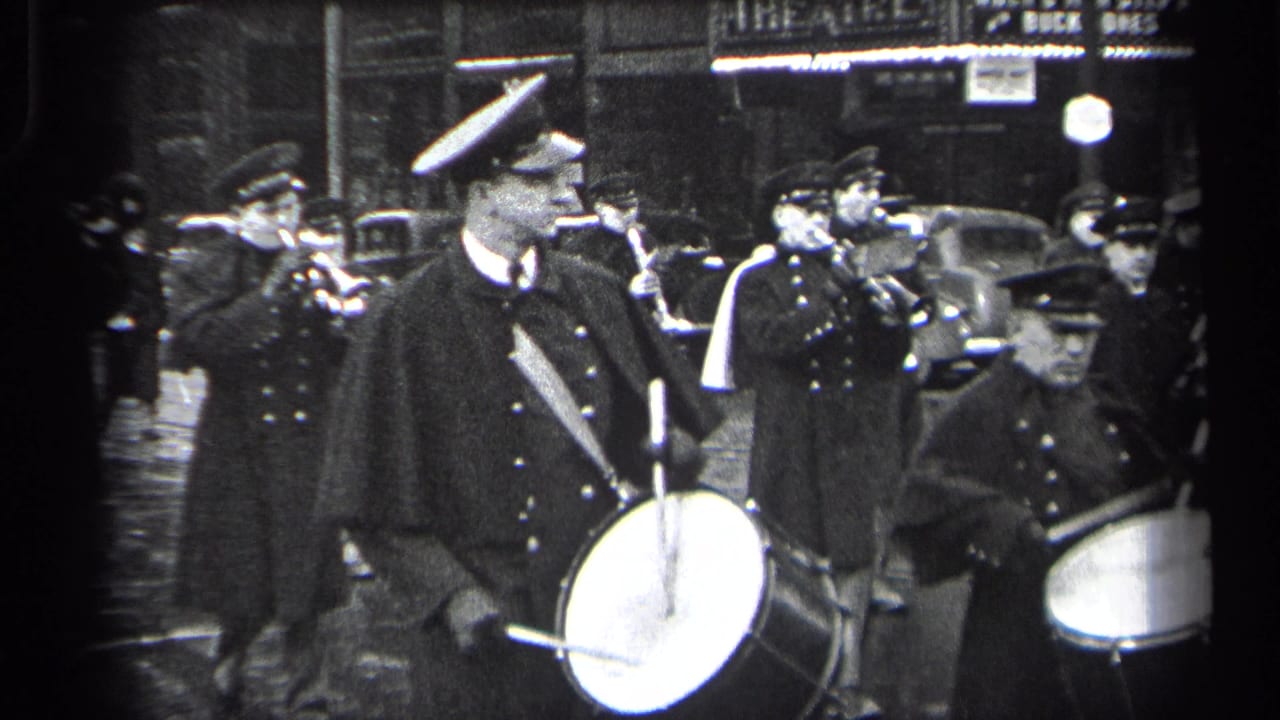 Drummers and brass players in dark uniforms march past cars and storefronts.