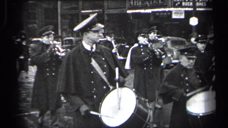 Uniformed musicians in Cleveland, Ohio, march at night with drums and brass instruments beside 1930s automobiles.