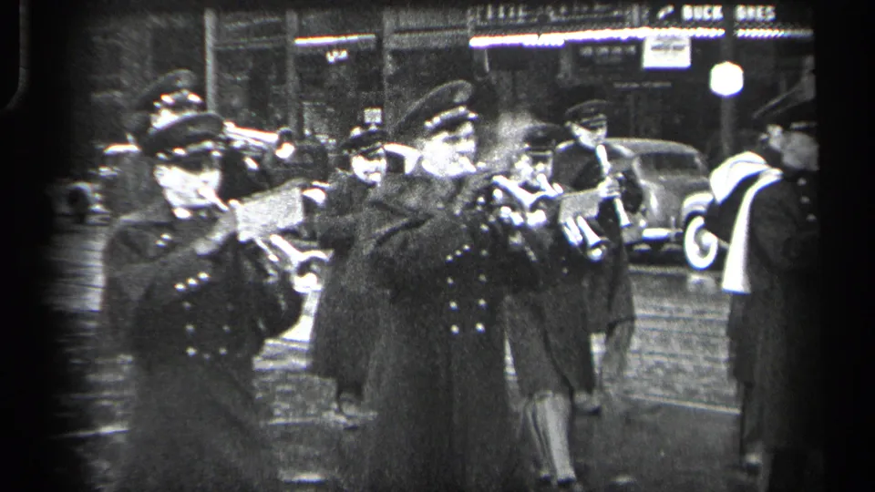 Two uniformed brass musicians play in a 1937 Cleveland nighttime procession with pedestrians and cars in the background.