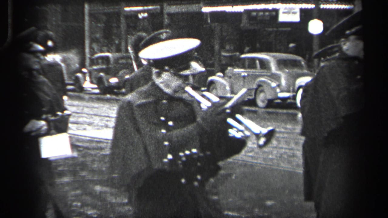 1937 Cleveland home-movie frame showing a uniformed parade participant with instruments near parked 1930s cars on a wet street.