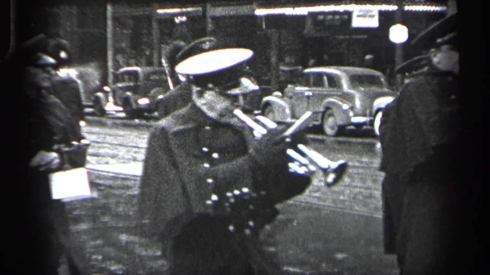1937 Cleveland home-movie frame showing a uniformed parade participant with instruments near parked 1930s cars on a wet street.