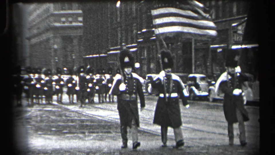 1937 Cleveland home-movie frame showing a flag bearer and uniformed marchers moving through a downtown street parade.