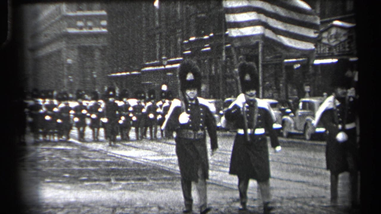 Uniformed marchers in tall hats walk toward the camera on a city street.
