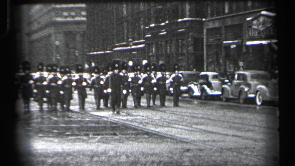 A larger uniformed formation moves along a Cleveland street, with 1930s automobiles parked at the curb and storefronts behind them.