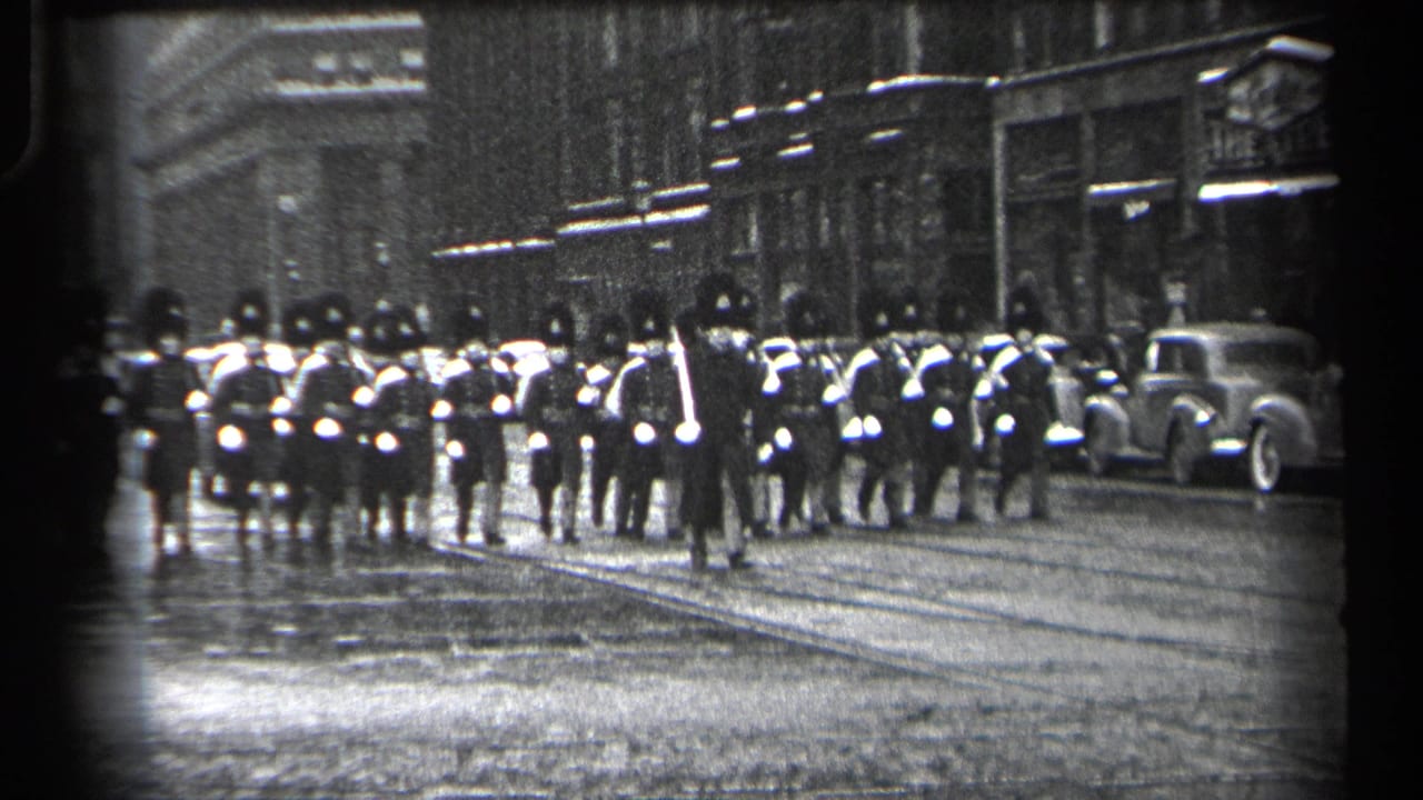 1937 Cleveland street parade with uniformed marchers in tall hats moving along wet pavement past storefronts and parked cars.