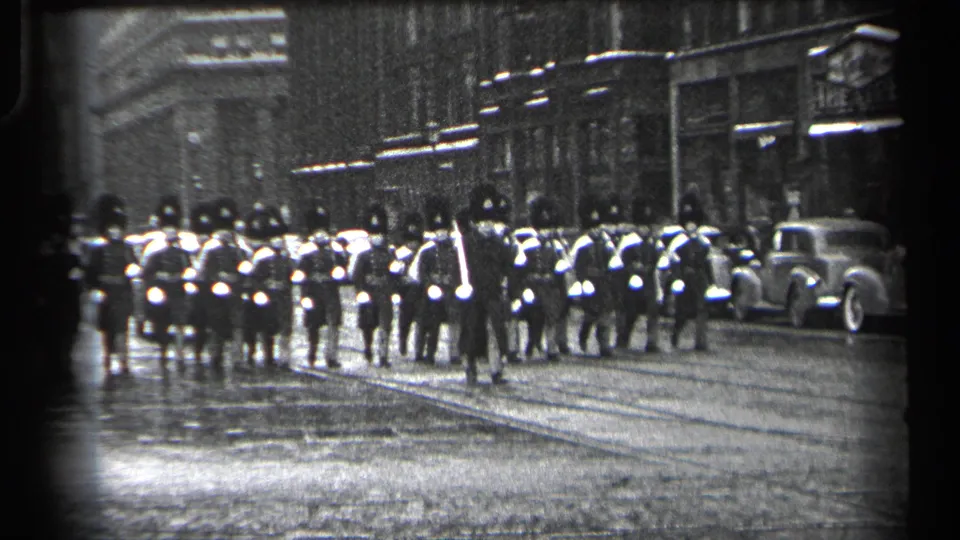 1937 Cleveland street parade with uniformed marchers in tall hats moving along wet pavement past storefronts and parked cars.