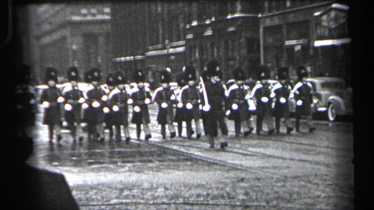 A parade formation crosses a wet street while a vintage car waits near storefronts.