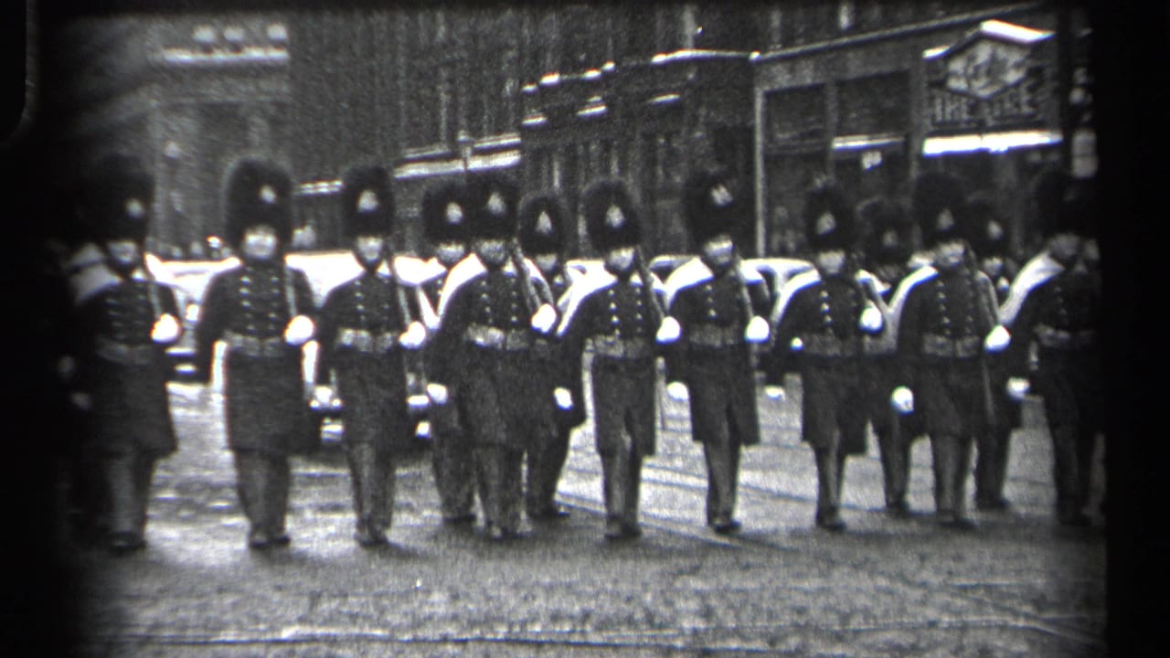 Marching guards in dark uniforms approach the camera with cars and shopfronts in the background.