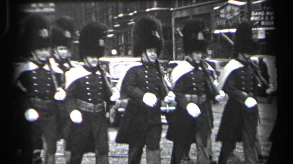 In 1937 Cleveland, ceremonial guards in tall bearskin-style hats and dark coats march past parked cars on a city street.