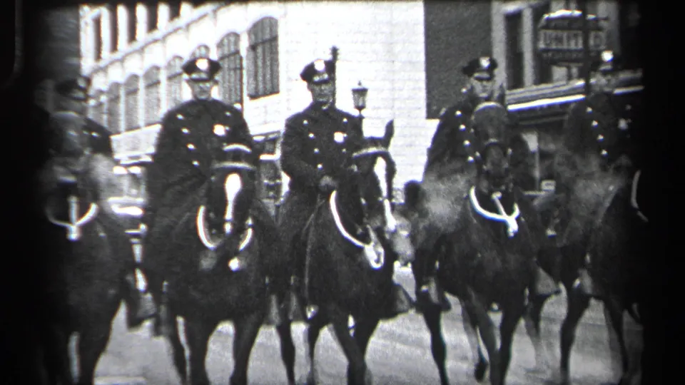 Mounted police in uniform ride horses past city buildings and street fixtures in this 1937 Cleveland 8mm frame.