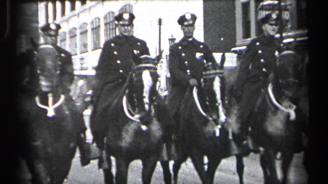Mounted policemen in dark uniforms ride horses along a Cleveland street in 1937, with buildings and traffic visible behind them.