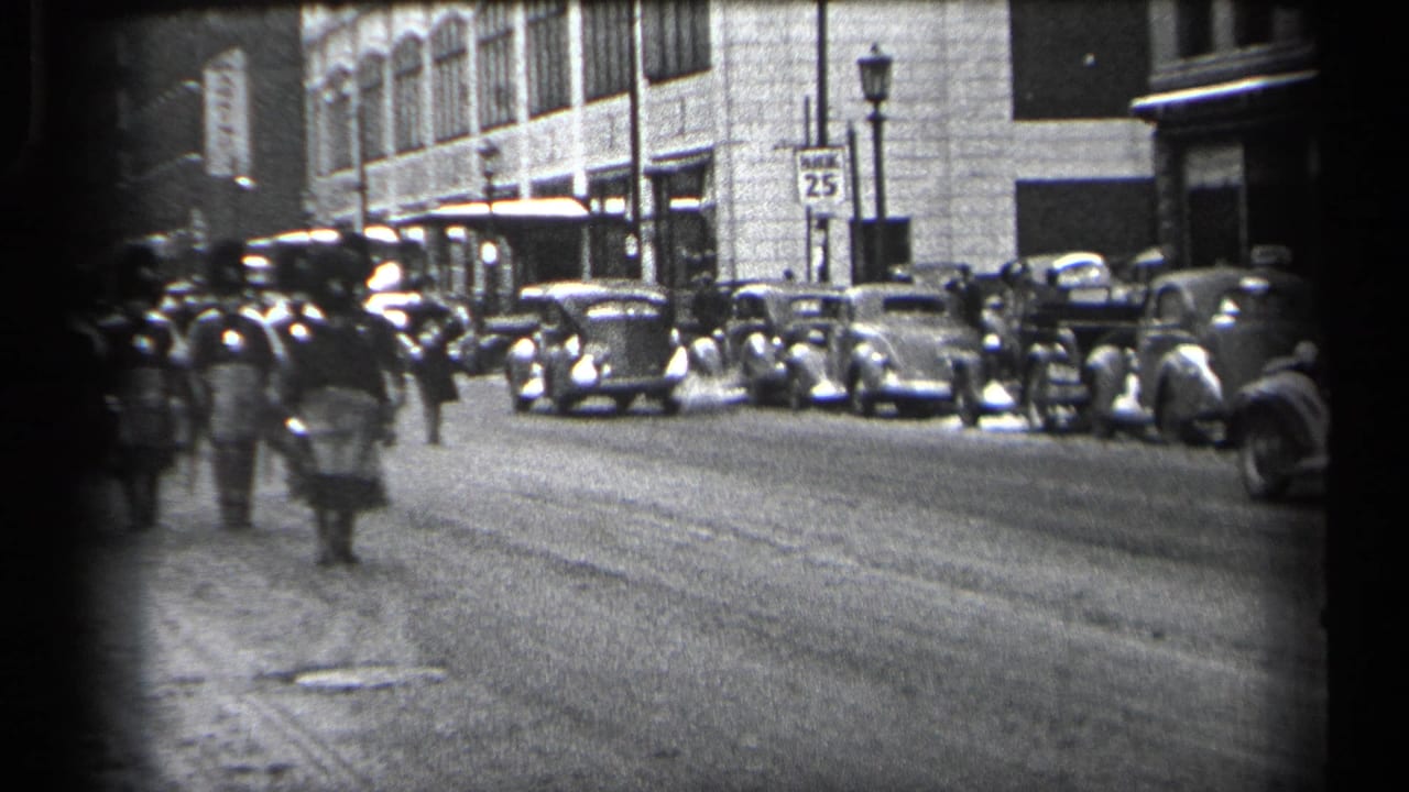 Uniformed marchers walk down a city street past parked 1930s cars.