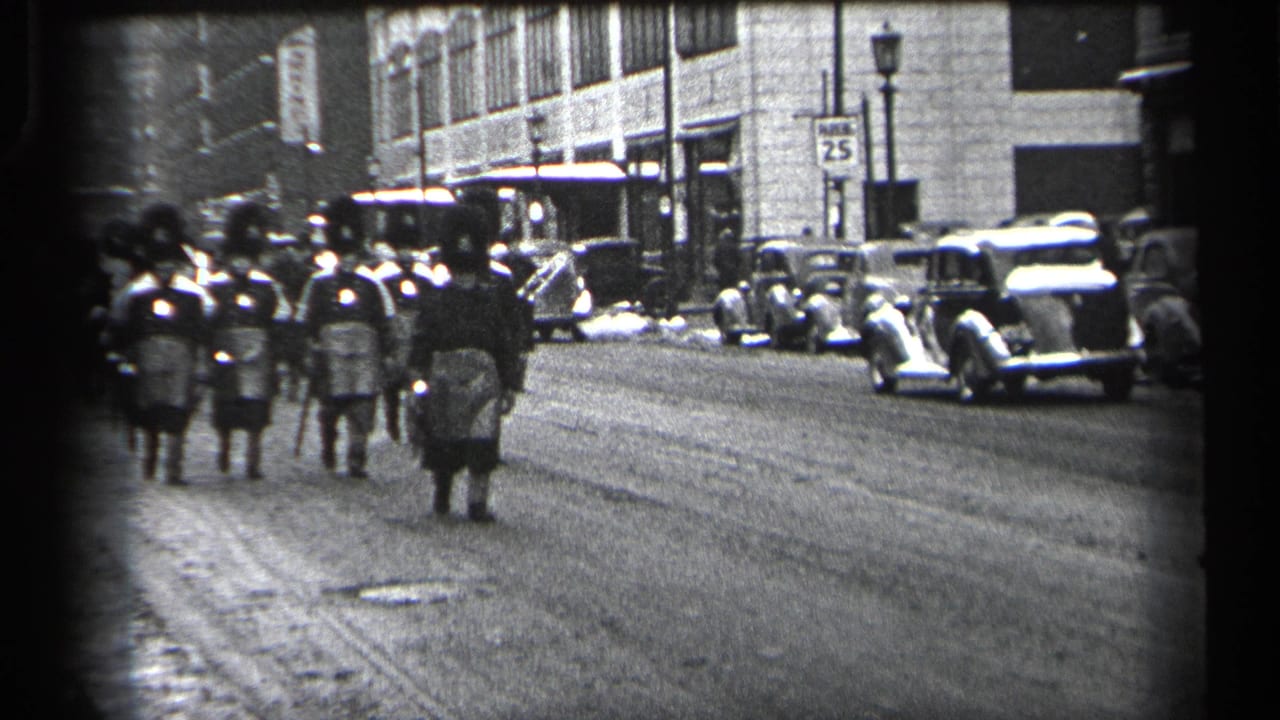 Marchers in tall hats and kilts proceed beside a row of parked cars.