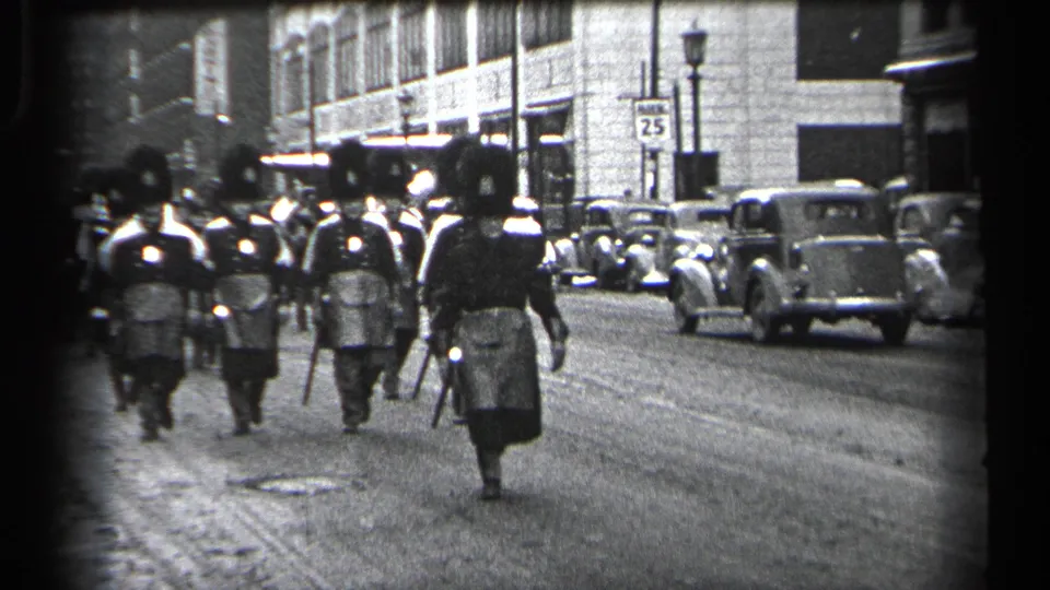 Uniformed marchers continue along the Cleveland street in 1937, carrying rifles past parked cars, storefront buildings, and a visible street sign.