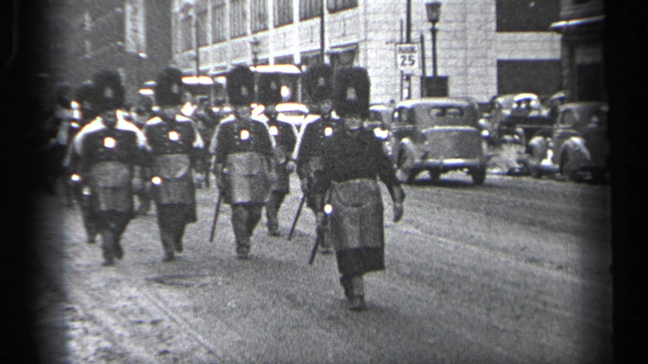 In Cleveland, Ohio, 1937, men in formal parade uniforms with tall hats and batons approach along a city street lined with cars.