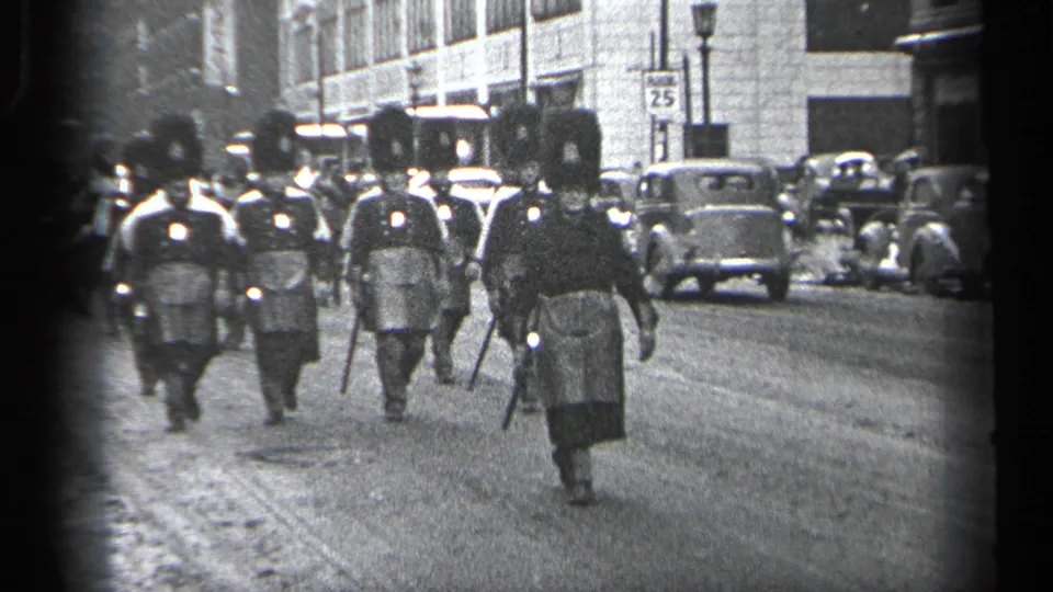 In Cleveland, Ohio, 1937, men in formal parade uniforms with tall hats and batons approach along a city street lined with cars.