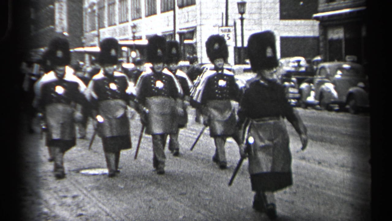 A line of uniformed parade marchers approaches the camera on a street.
