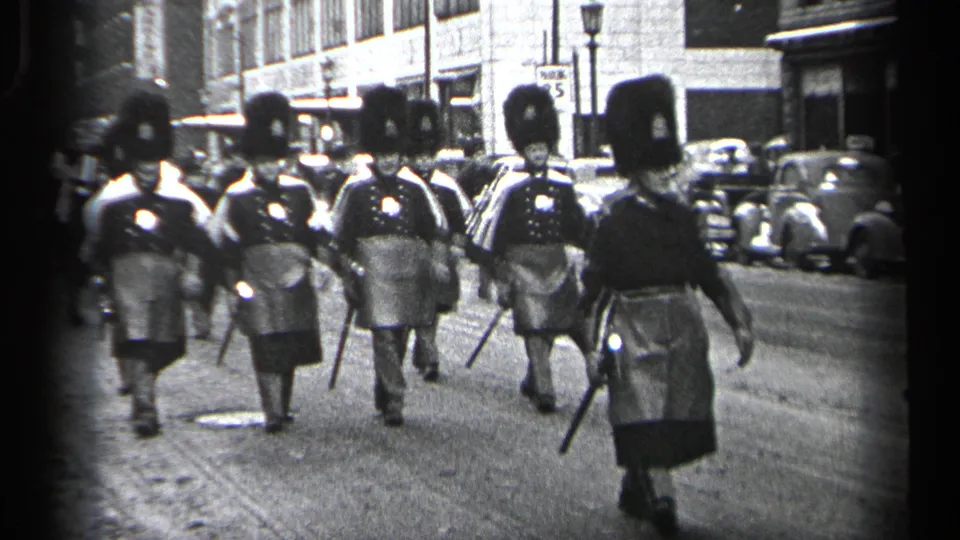Men in formal uniforms with tall hats and batons march in formation on a Cleveland street, with 1930s cars visible in the background.