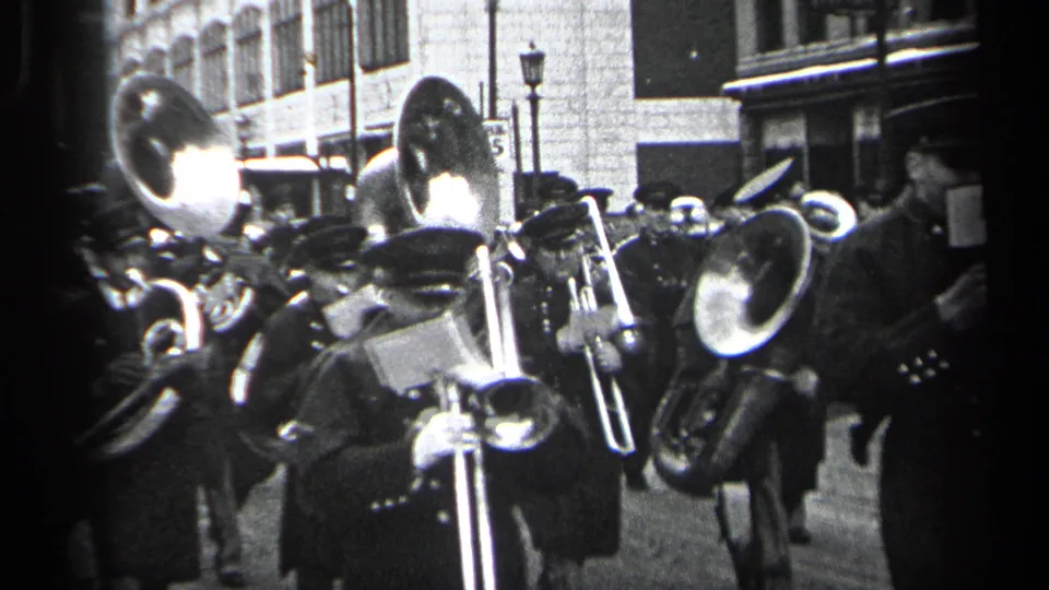 In 1937 Cleveland, uniformed marching band members pass close to the camera with trombones and large brass horns visible.