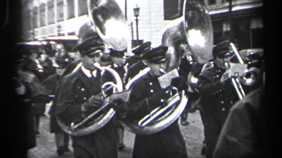 Uniformed musicians in the Cleveland parade carry curved brass horns and sheet music while spectators line the sidewalk.