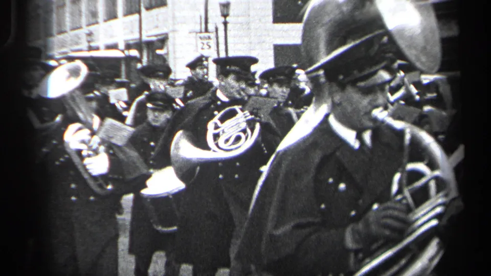 A close view shows a uniformed brass player in front, with other Cleveland parade musicians and a 25 speed limit sign behind him.