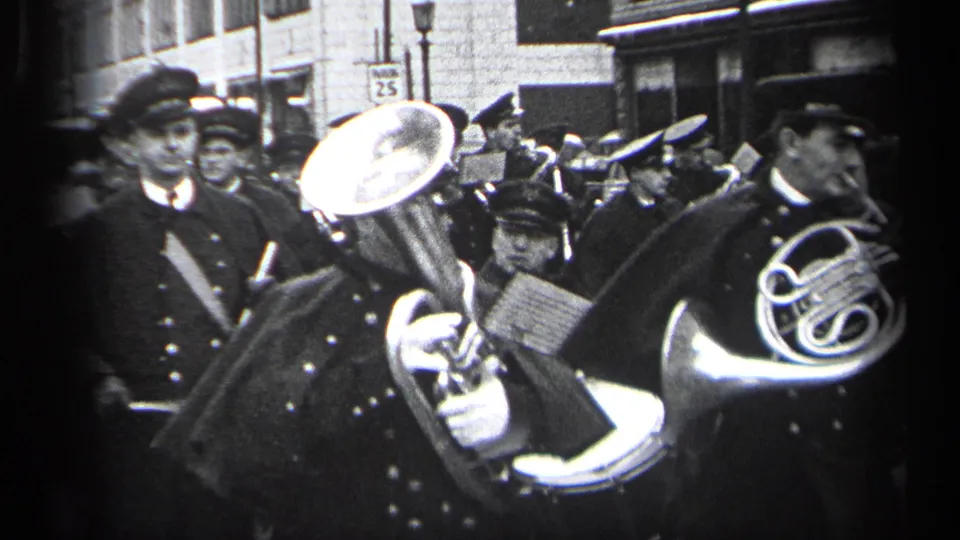 Band members in dark uniforms march through Cleveland with brass instruments, sheet music, spectators, and a 25 speed limit sign visible.