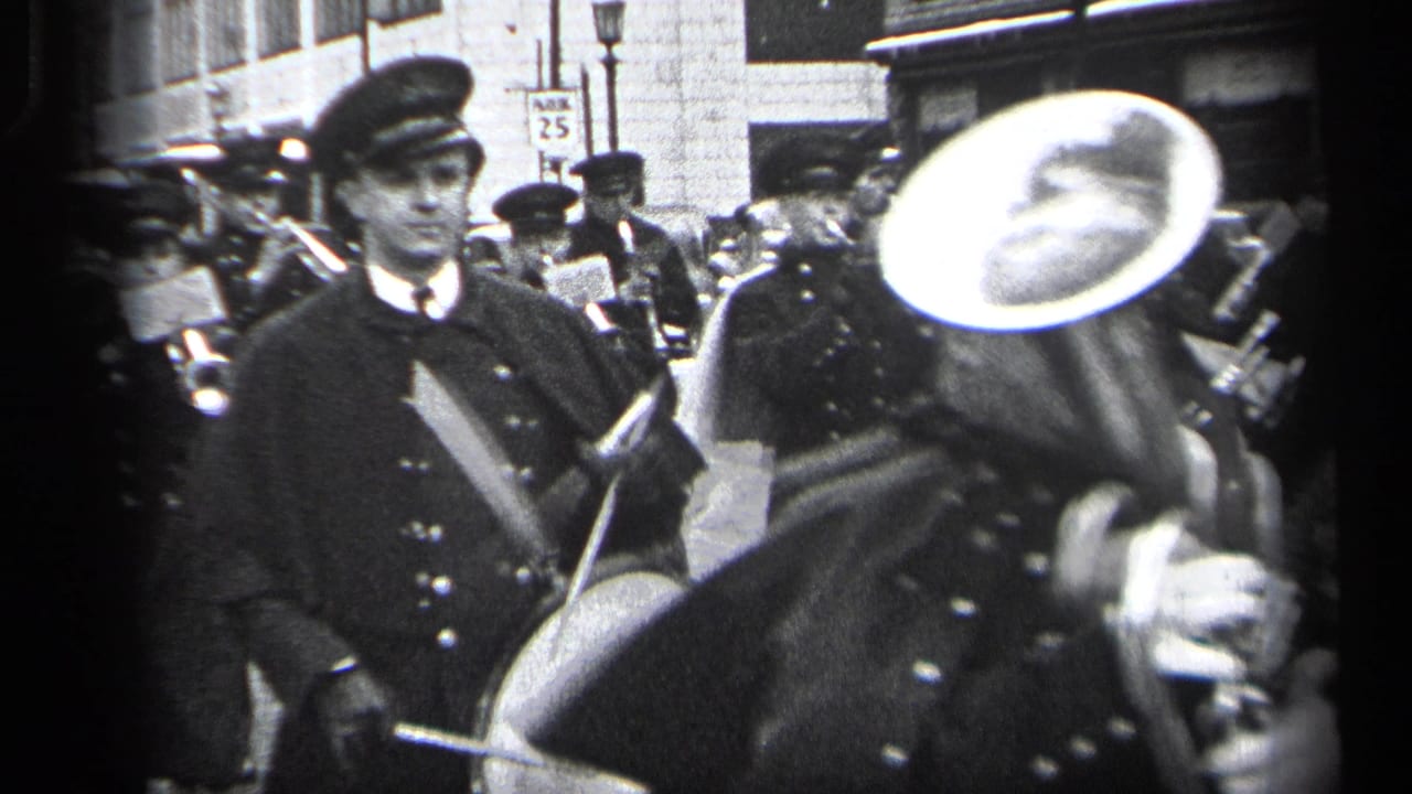 Uniformed marchers and band members pass on a city street, with a large tuba bell near the camera.