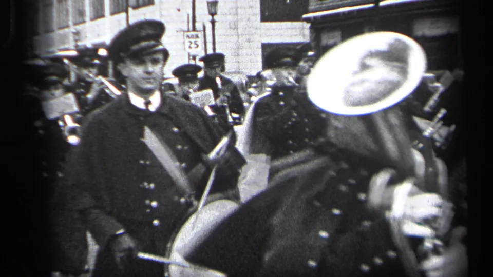 1937 Cleveland parade frame showing uniformed adults in peaked caps marching past the camera with brass instruments on a city street.