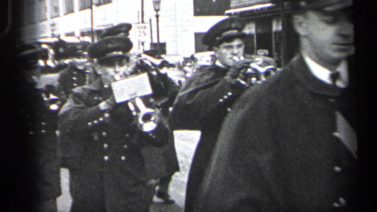 Uniformed band members with trumpets march past buildings and automobiles in the background.