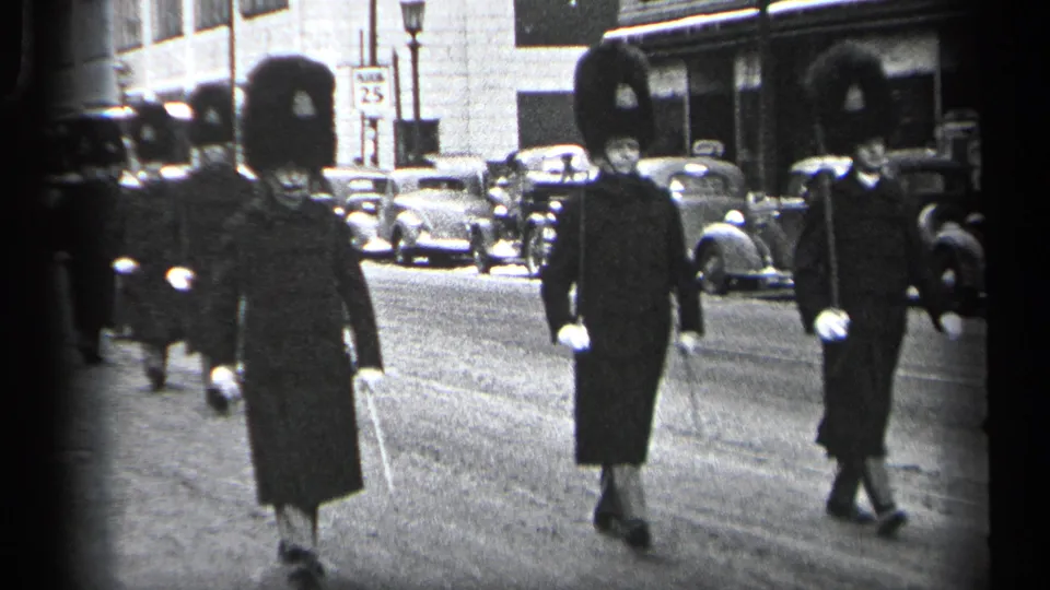 Uniformed guards in tall bearskin-style hats march down a Cleveland street lined with 1930s cars.
