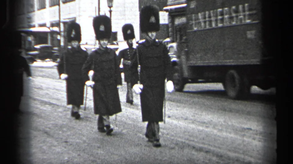 In 1937 Cleveland, uniformed guards in tall dark hats march along a city street past parked and moving vehicles.