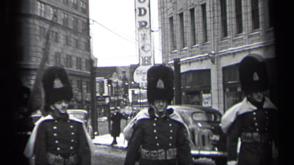 1937 Cleveland street parade with uniformed marchers in tall fur hats passing storefronts, parked cars, and a vertical sign.