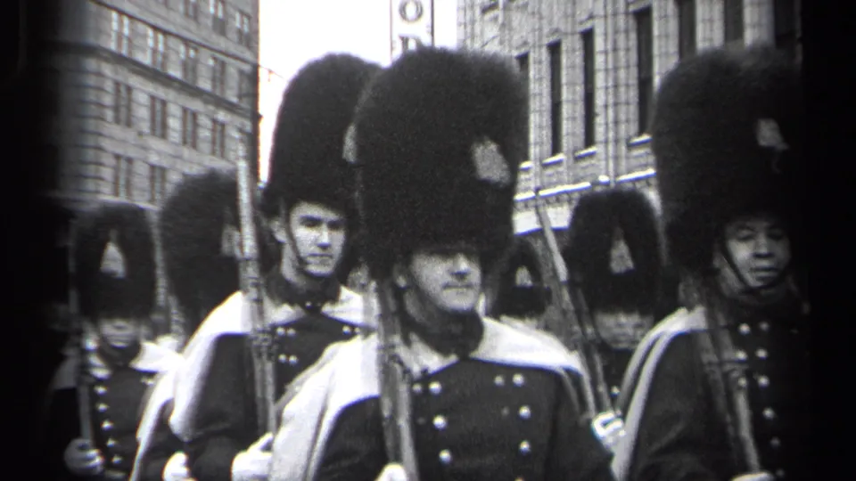 1937 Cleveland home-movie frame showing uniformed men in tall bearskin-style hats marching with rifles past urban buildings.