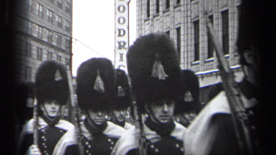Uniformed men in tall black hats and caped dress uniforms march in formation on a Cleveland street in 1937 footage.