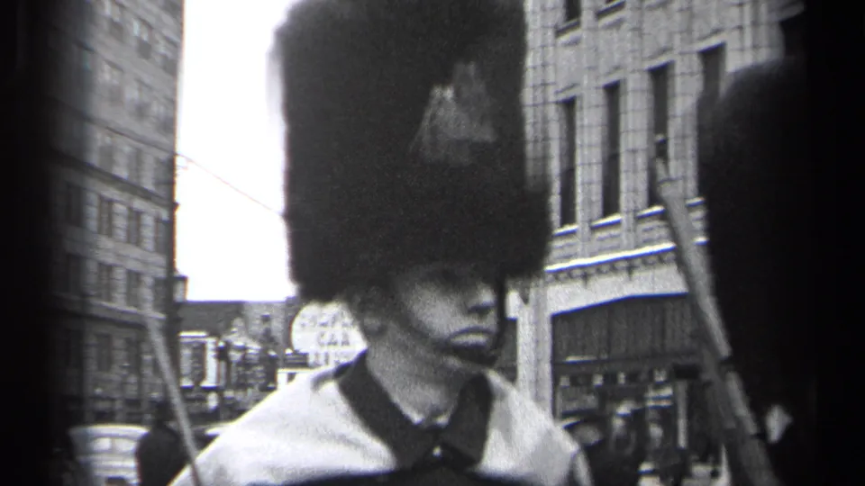 Close 1937 Cleveland frame of a uniformed parade marcher in a tall dark hat, with rifle and downtown buildings visible nearby.
