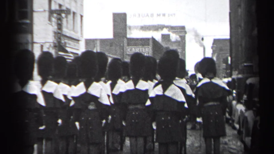Uniformed parade marchers in tall hats continue through a Cleveland street scene with storefronts and parked 1930s automobiles.