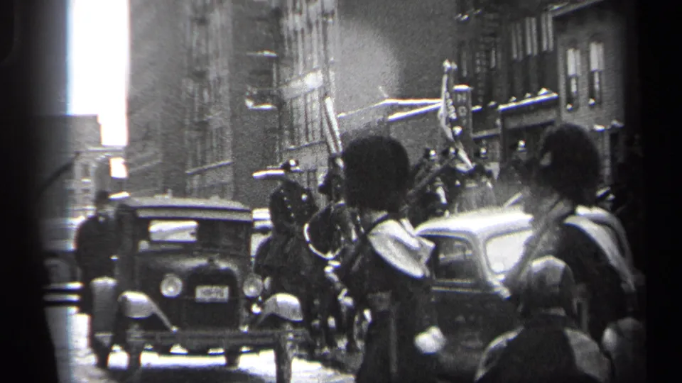 Street-level 1937 Cleveland parade frame showing marchers, a visible standard, spectators, and prewar automobiles in the roadway.