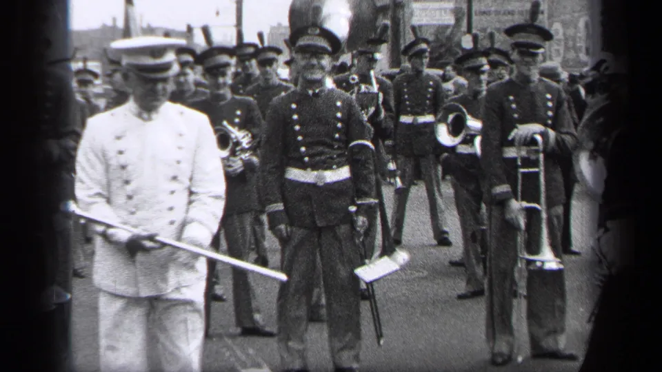 The baton leader faces forward in 1937 Cleveland as uniformed musicians with trombone and brass instruments stand behind him.