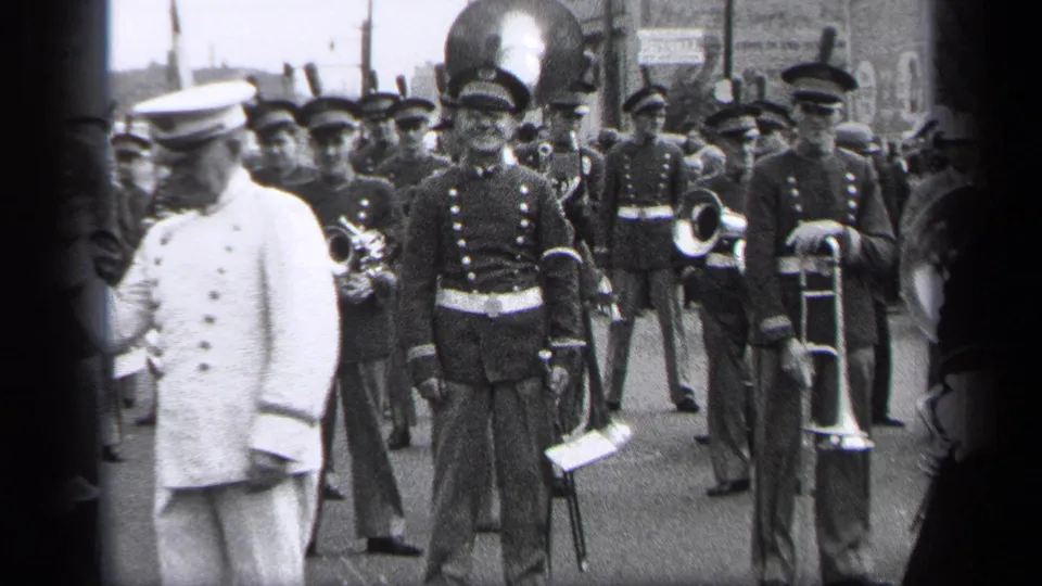 In the Cleveland parade scene, the white-uniformed leader turns left while dark-uniformed musicians with brass instruments fill the frame.