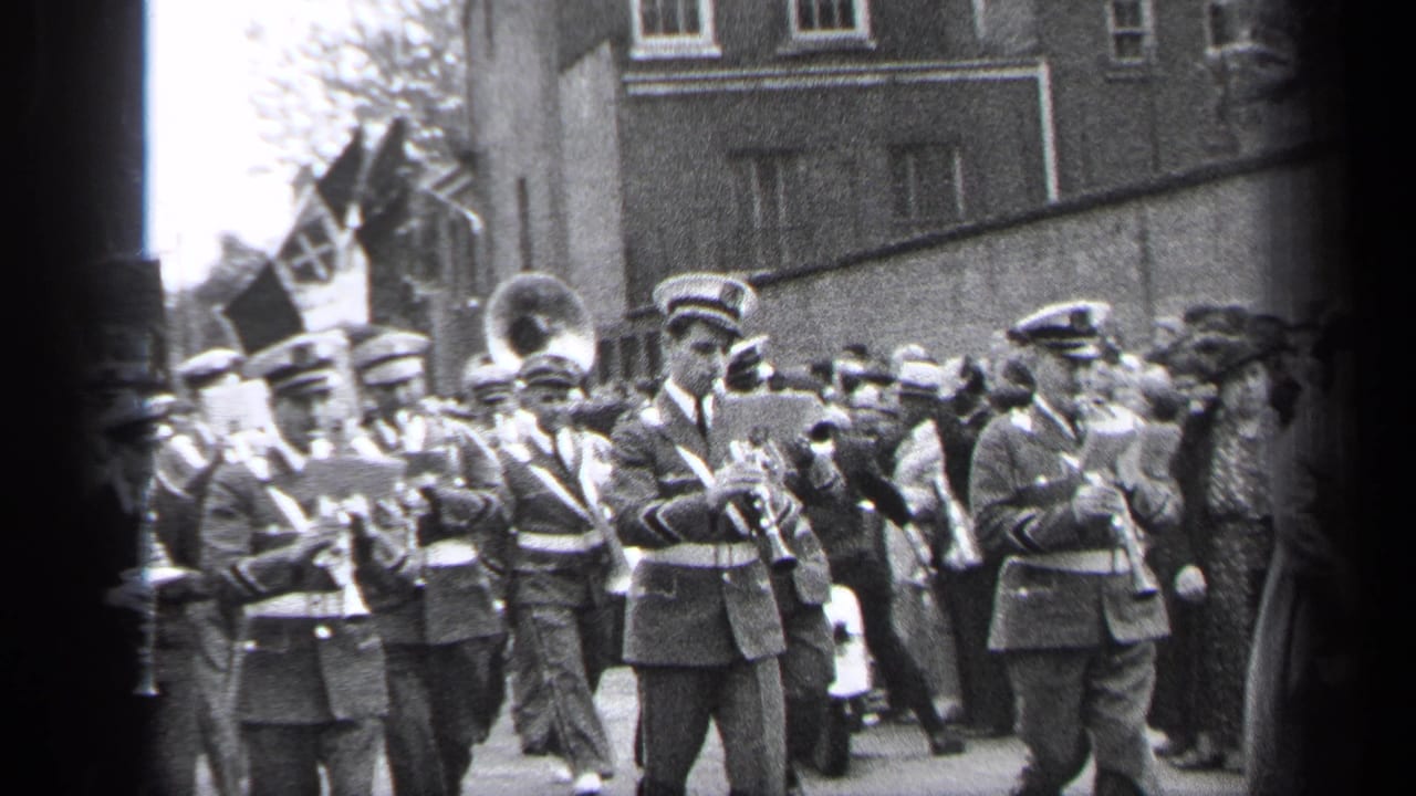 In 1937 Cleveland, uniformed band members march past a crowd with flags and residential buildings behind them.