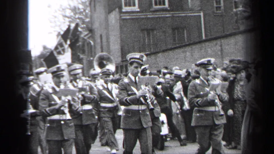 In 1937 Cleveland, uniformed band members march past a crowd with flags and residential buildings behind them.