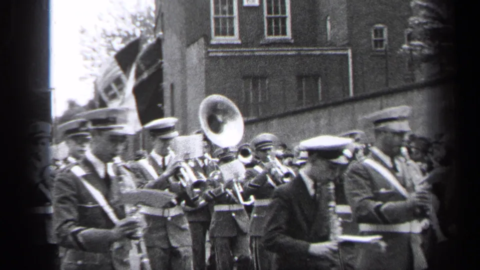 A 1937 Cleveland parade frame shows uniformed band members, a prominent tuba or sousaphone bell, flags, and onlookers near a city building.