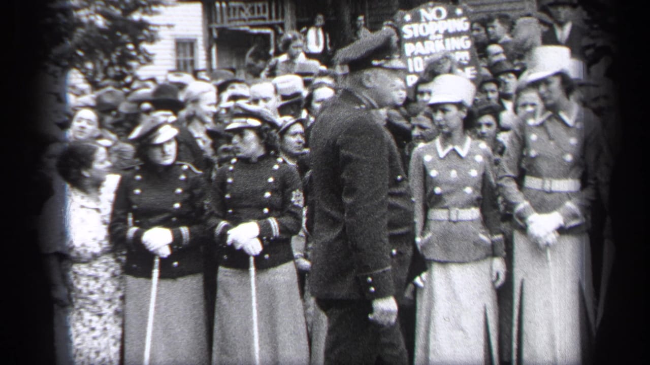 Uniformed women with white gloves and canes stand before a crowd while a man faces them.