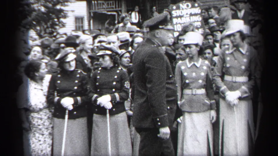 In 1937 Cleveland, uniformed women with caps, white gloves, and canes stand in formation as a man addresses them before onlookers.