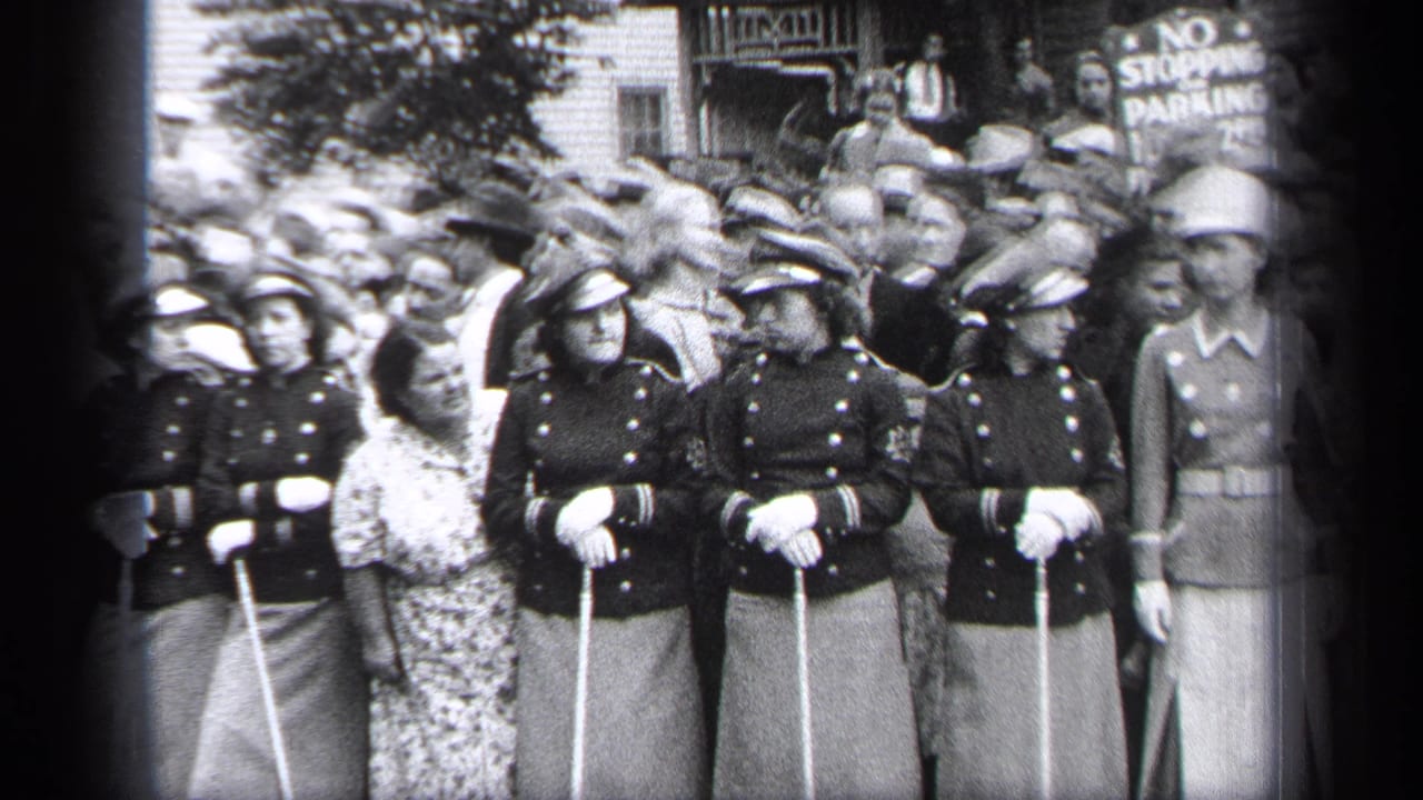 Women in dark uniforms hold canes in front of a packed crowd and street sign.