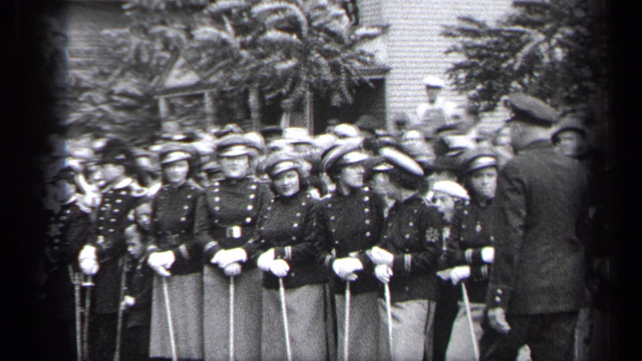 A man in a cap walks past uniformed young women standing in formation.