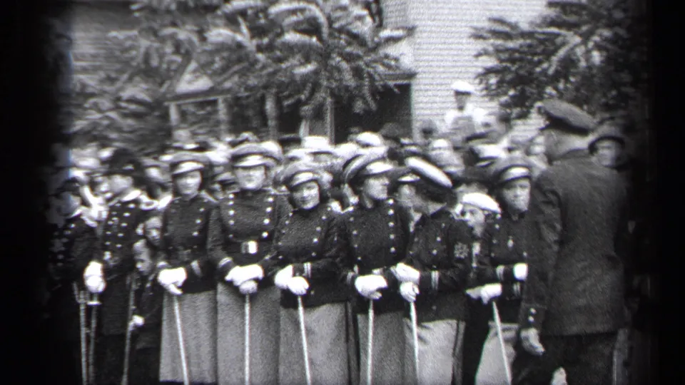 In the 1937 Cleveland footage, a capped man moves in front of young women in matching uniforms as spectators stand behind them.