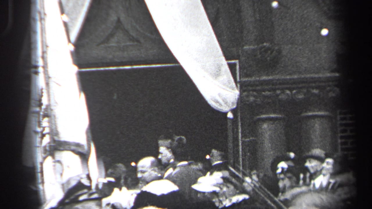 Crowd in hats gathered near a large flag and draped fabric outside dark building doors.
