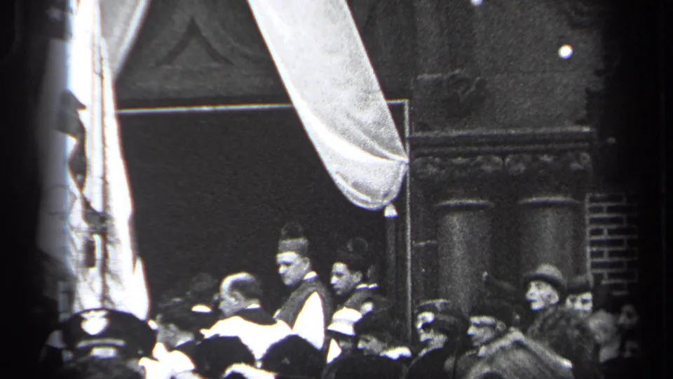 1937 Cleveland home-movie frame with a crowd in period dress near a large flag, white drape, and dark building entrance.